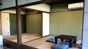 Une pièce avec un sol recouvert de tatamis, une petite table et des coussins, des poutres en bois et un climatiseur fixé au mur.