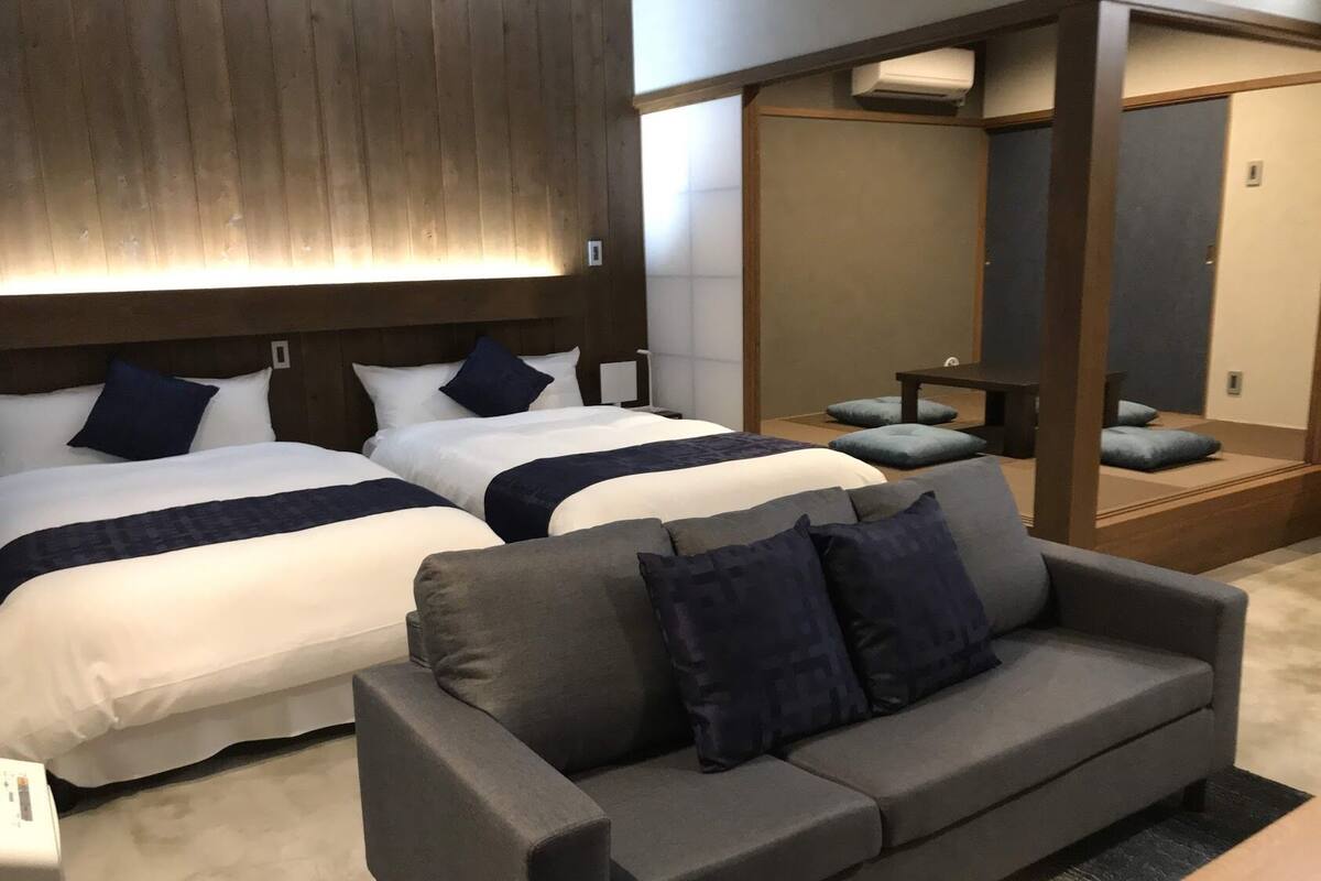 Quarto de hotel com duas camas, um sofá cinza e uma cabeceira de madeira.