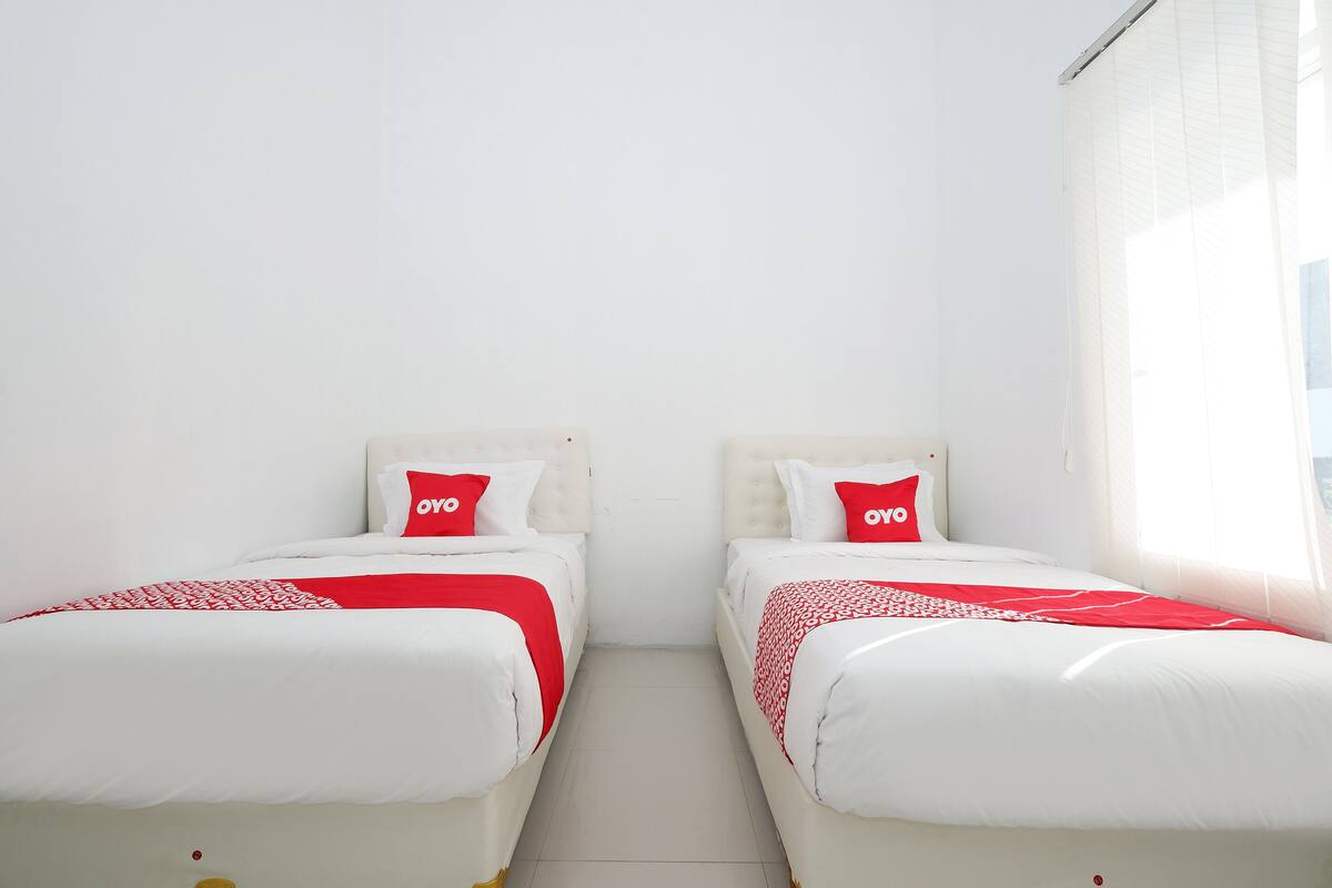 Habitación de hotel con dos camas, cada una con ropa blanca y detalles rojos, frente a una pared blanca lisa.