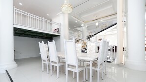 Un comedor espacioso con sillas blancas y una mesa de mármol, rodeado de columnas blancas y una escalera que conduce a un nivel superior.