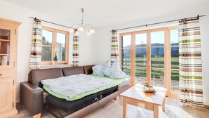 A room with a sofa bed, a table, and a view of a green landscape through large windows. - Cosy Apartment "Bergblick" with Garden, Terrace & Wi-Fi; Parking Available (Bad Heilbrunn)