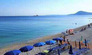 Une plage avec une eau d’un bleu limpide, un rivage rocheux, ainsi que plusieurs parasols et chaises longues.