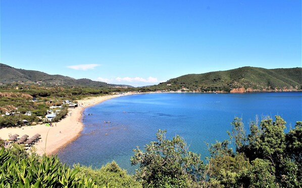 Un paysage côtier avec une plage, une eau d’un bleu limpide et des collines en arrière-plan.