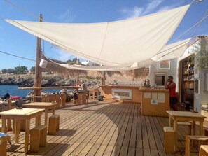 A wooden deck with tables and chairs, a bar area, and a view of the sea.