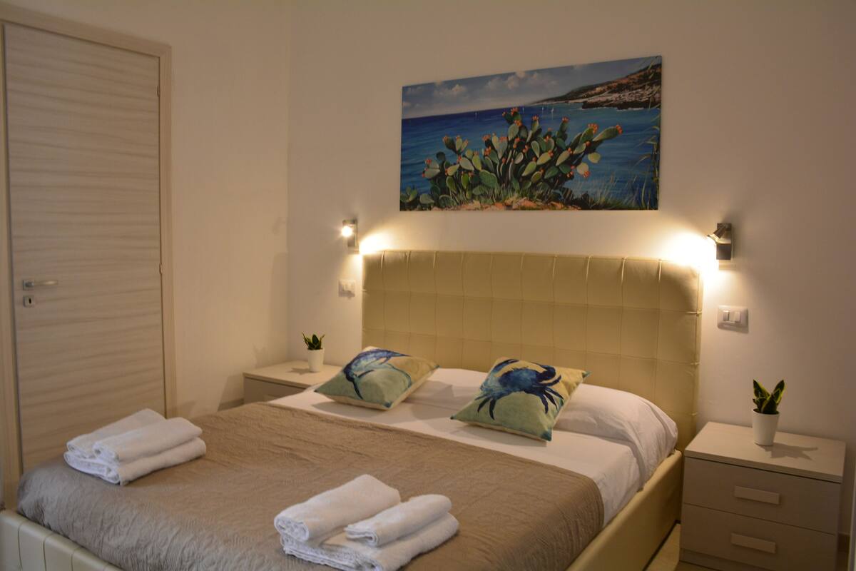 A neatly made bed with towels, a headboard with lights, a painting of a cactus and ocean, and a closed door.