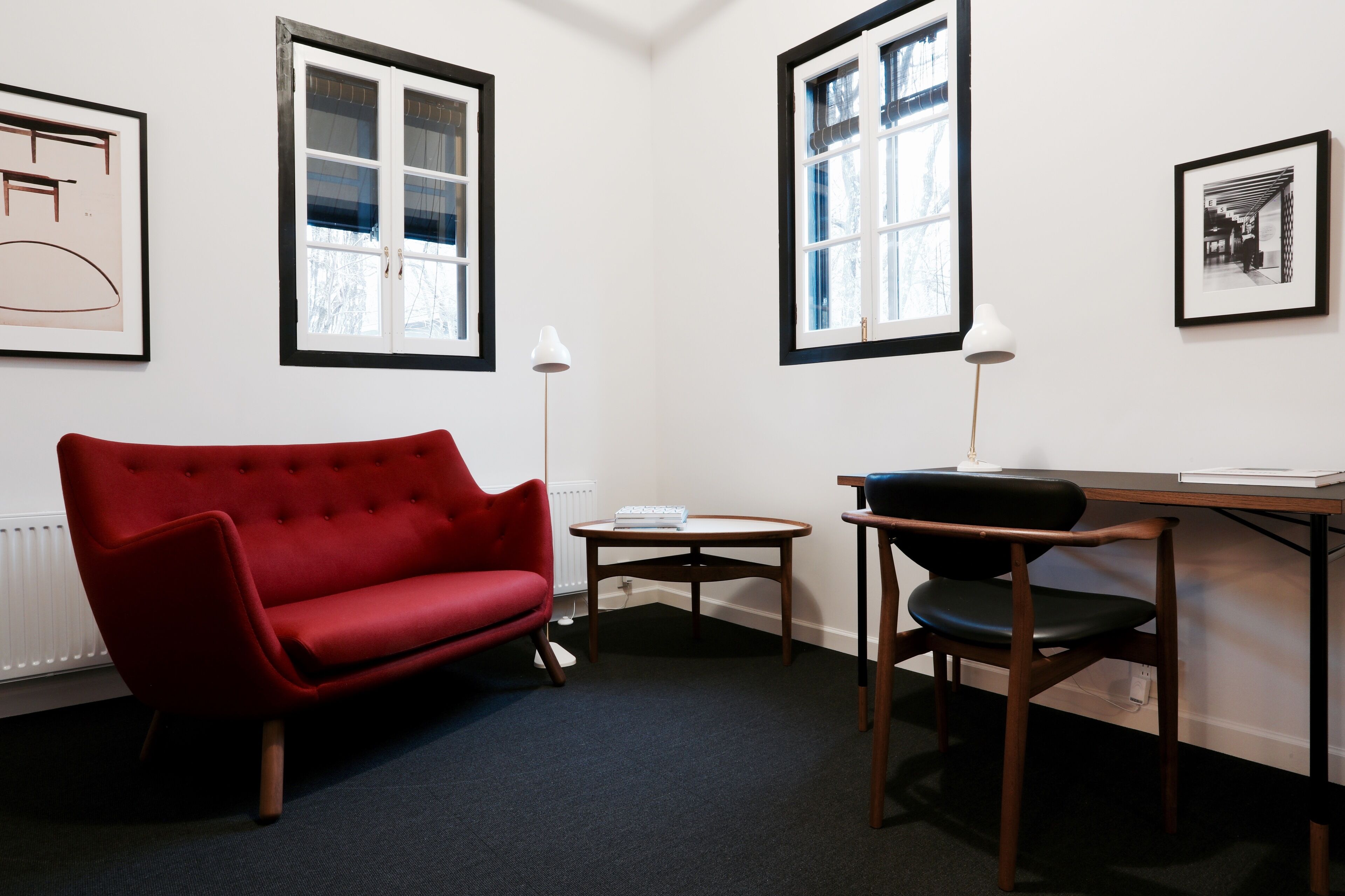 Ein Zimmer mit einem roten Sofa, einem Holztisch und zwei Stühlen. Es gibt zwei Fenster mit schwarzen Rahmen und weißem Zierstreifen.
