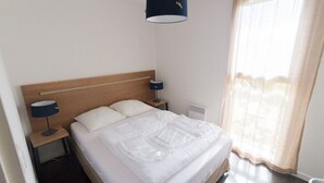 Habitación de hotel con cama, mesita de noche, lámpara, ventana con cortinas e iluminación en el techo.