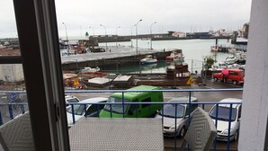 Ein Balkon mit Korbmöbeln, der auf einen Hafen mit Booten und einem roten Auto blickt.