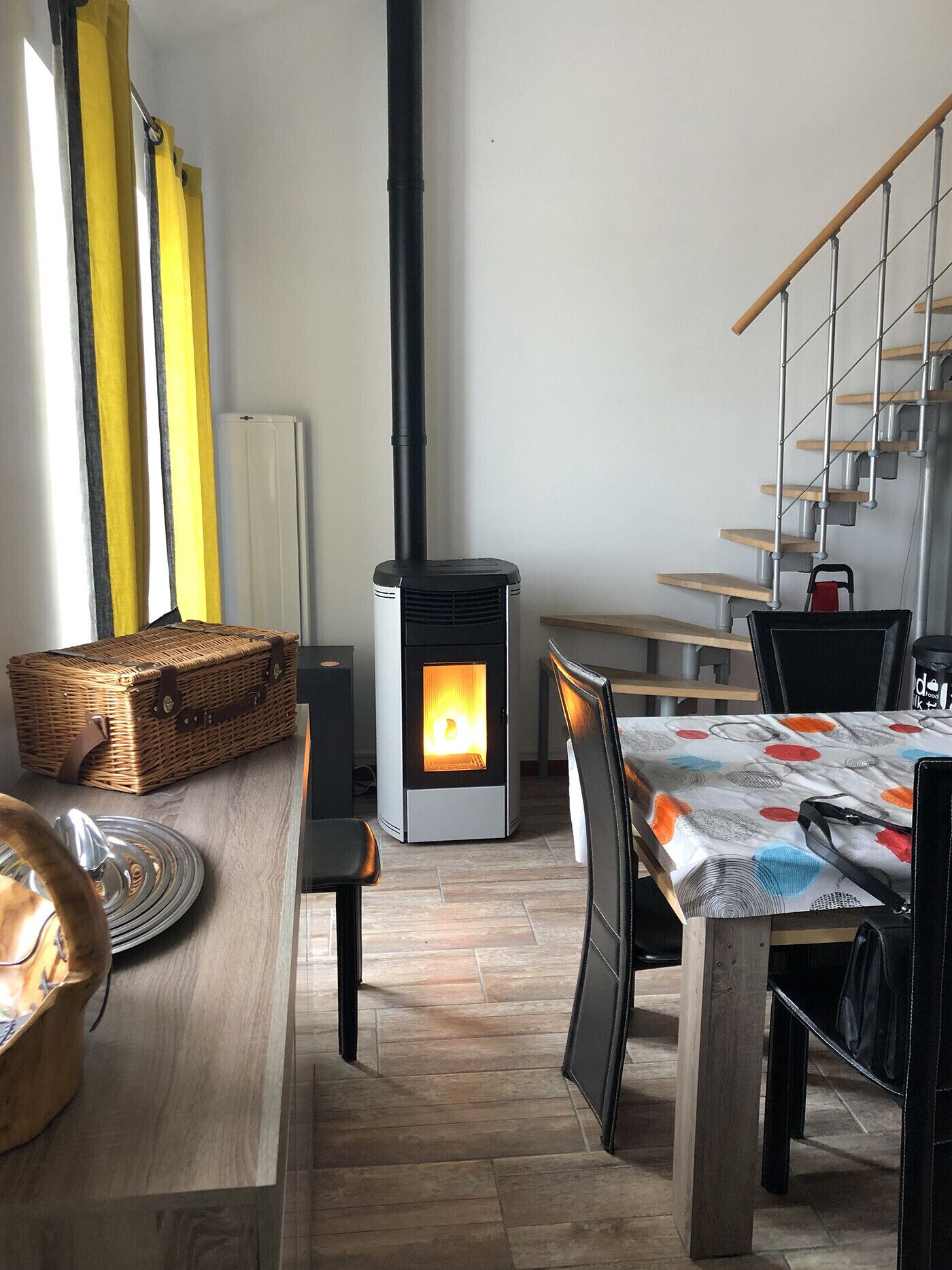 A cozy room with a wood-burning stove, a dining table, chairs, a wicker basket, and a staircase.