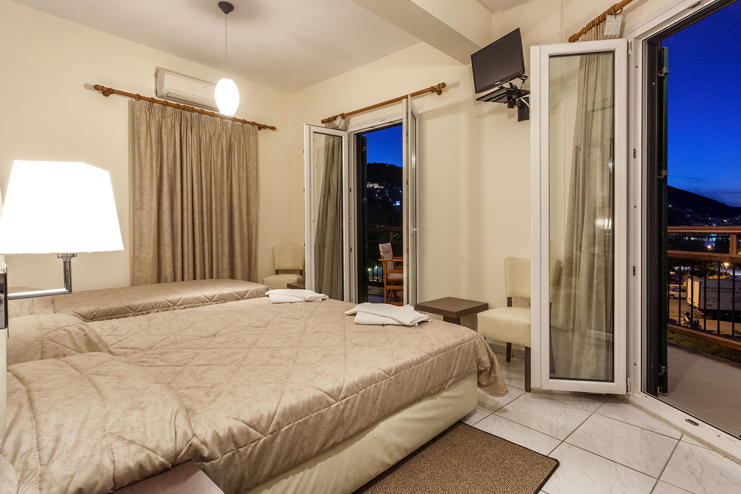 A hotel room with two beds, a sitting area, a TV, and a balcony with a city view.