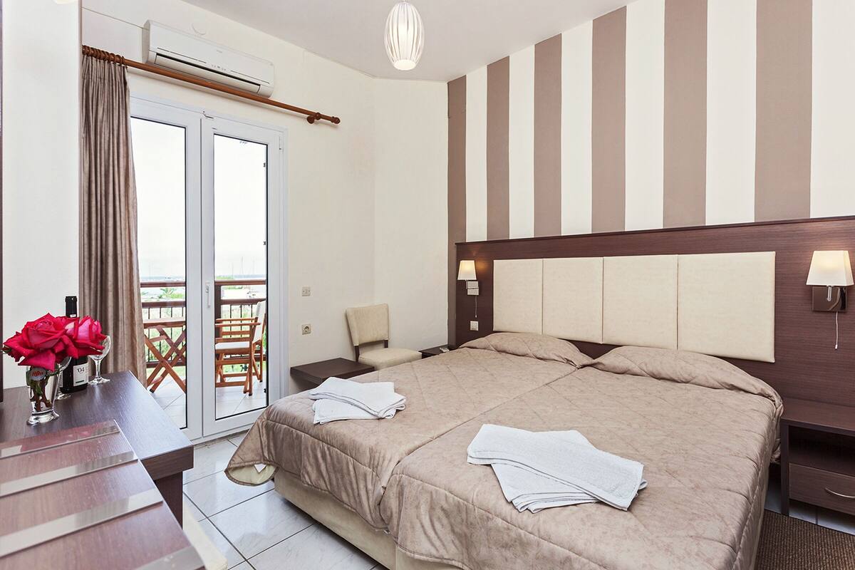 A hotel room with a large bed, a desk with a vase of flowers, a chair, and a view of the outdoors through a sliding door.