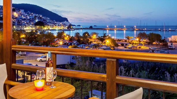 A balcony with a view of a coastal town at dusk, featuring a table with a bottle and glasses, and a stop sign in the foreground.