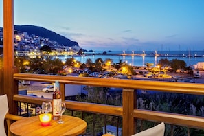Un balcón con vistas a un pueblo costero al anochecer, que incluye una mesa con una botella y vasos, y una señal de stop en primer plano.
