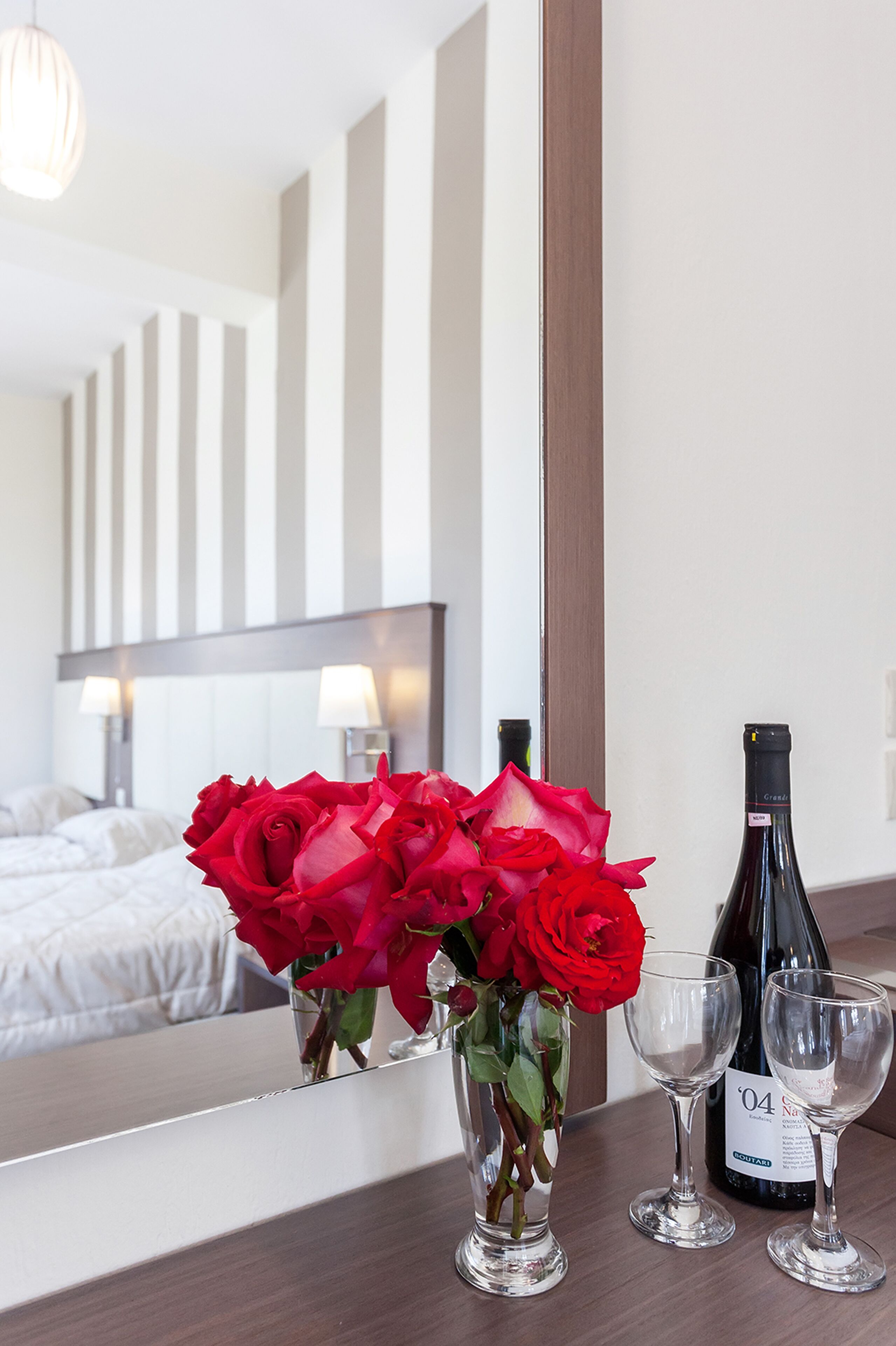 A hotel room with a bed, a vase with red roses, two wine glasses, and a bottle of wine on the nightstand.