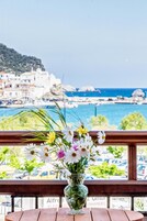 Una mesa de madera con un jarrón de cristal transparente que contiene flores blancas y rosas, con vistas a un pueblo costero con edificios y al mar.
