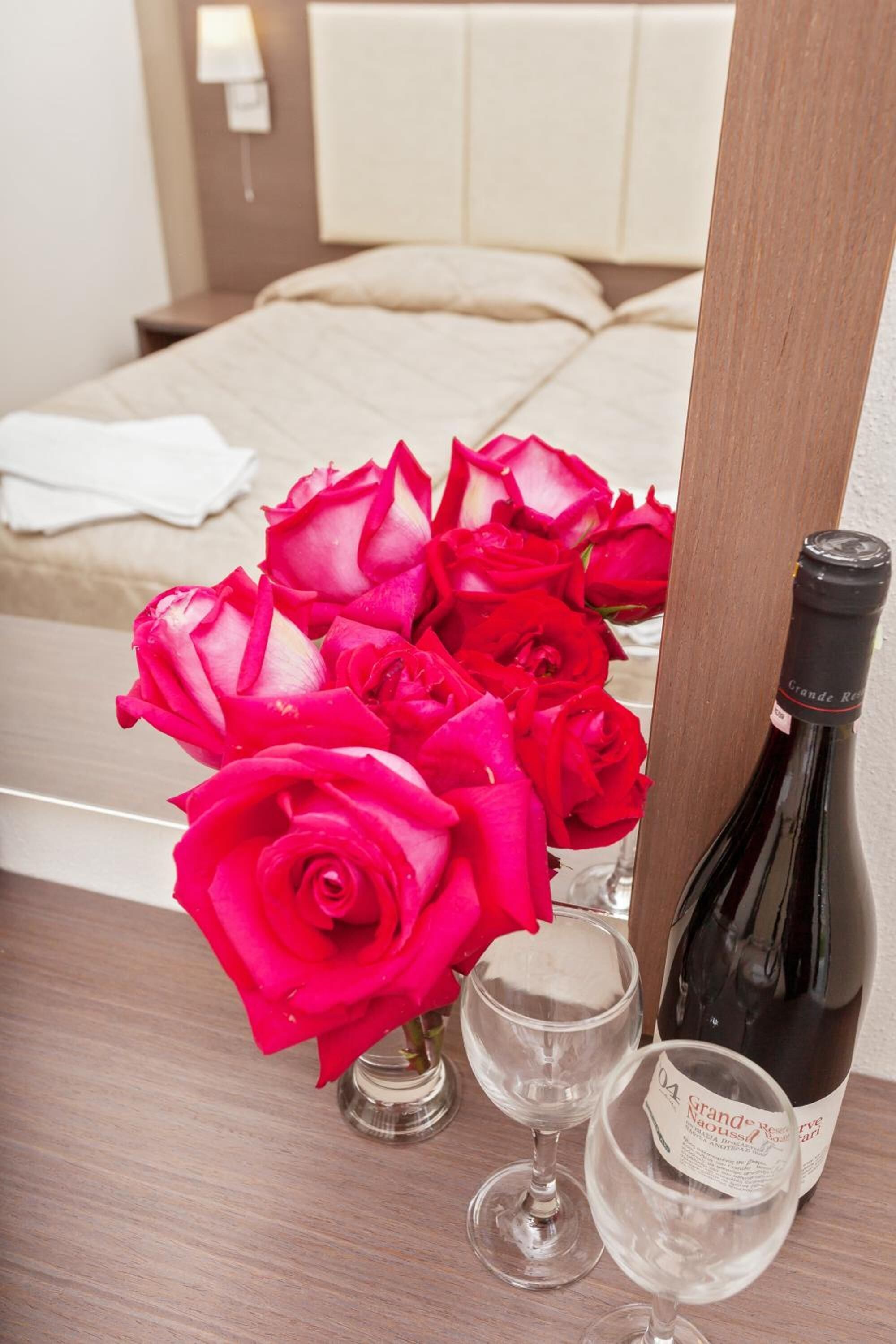 A hotel room with a bed, a nightstand, a bottle of wine, two wine glasses, and a vase with red roses.