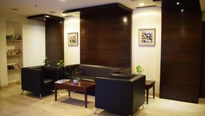 A modern lobby with a dark wood accent wall, a black leather sofa, two armchairs, a coffee table, and a bookshelf with books and decorative items. - Hotel Grenville (Gurugram)