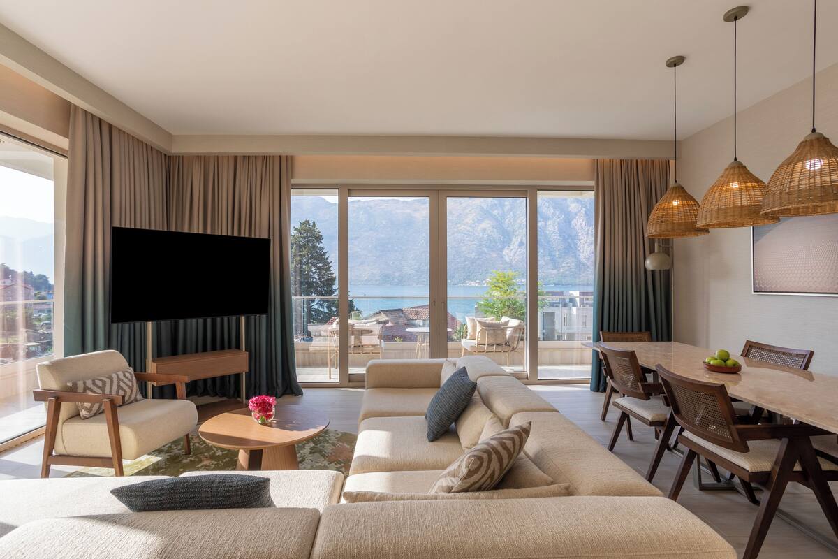 Un salon moderne doté d’un grand téléviseur à écran plat, d’un ensemble de canapés, d’une table à manger avec des chaises, et offrant une vue sur les montagnes grâce à de grandes fenêtres.