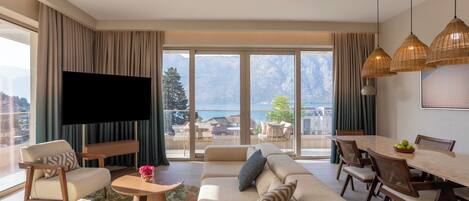 A modern living room with a large flat-screen TV, a sofa set, a dining table with chairs, and a view of mountains through large windows.