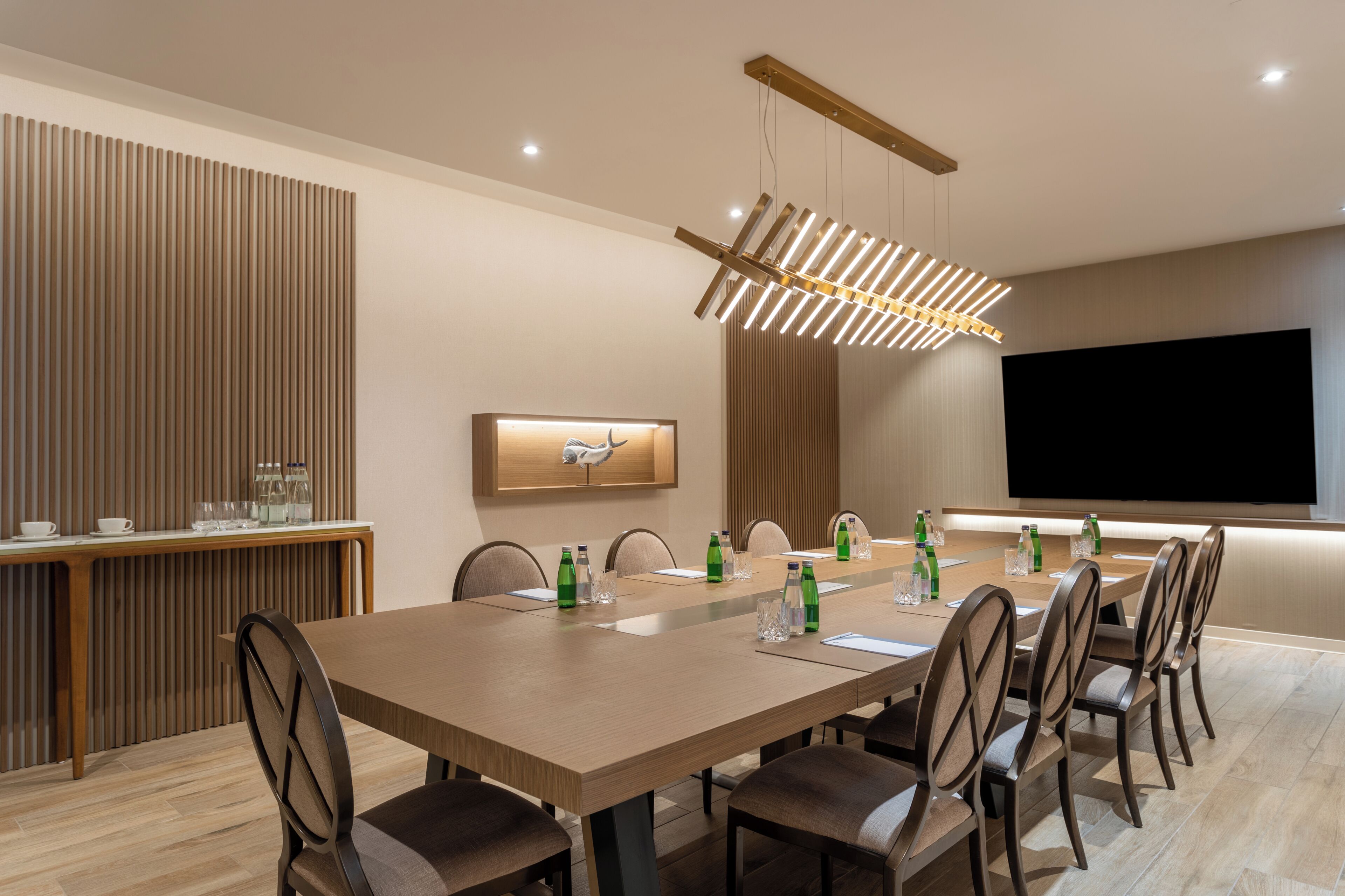 A modern dining area with a long table, chairs, a chandelier, a flat-screen TV, and a wooden accent wall.