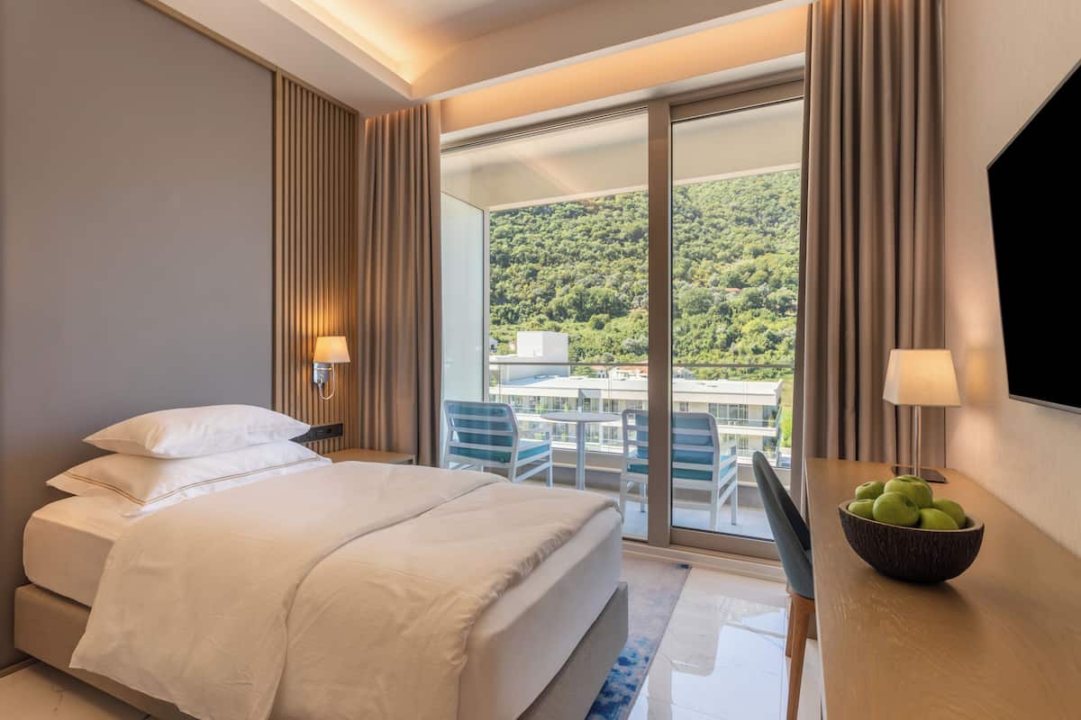 Une chambre d’hôtel avec un lit, un bureau sur lequel se trouve un panier de fruits, une télévision et un balcon offrant une vue sur une colline verdoyante.