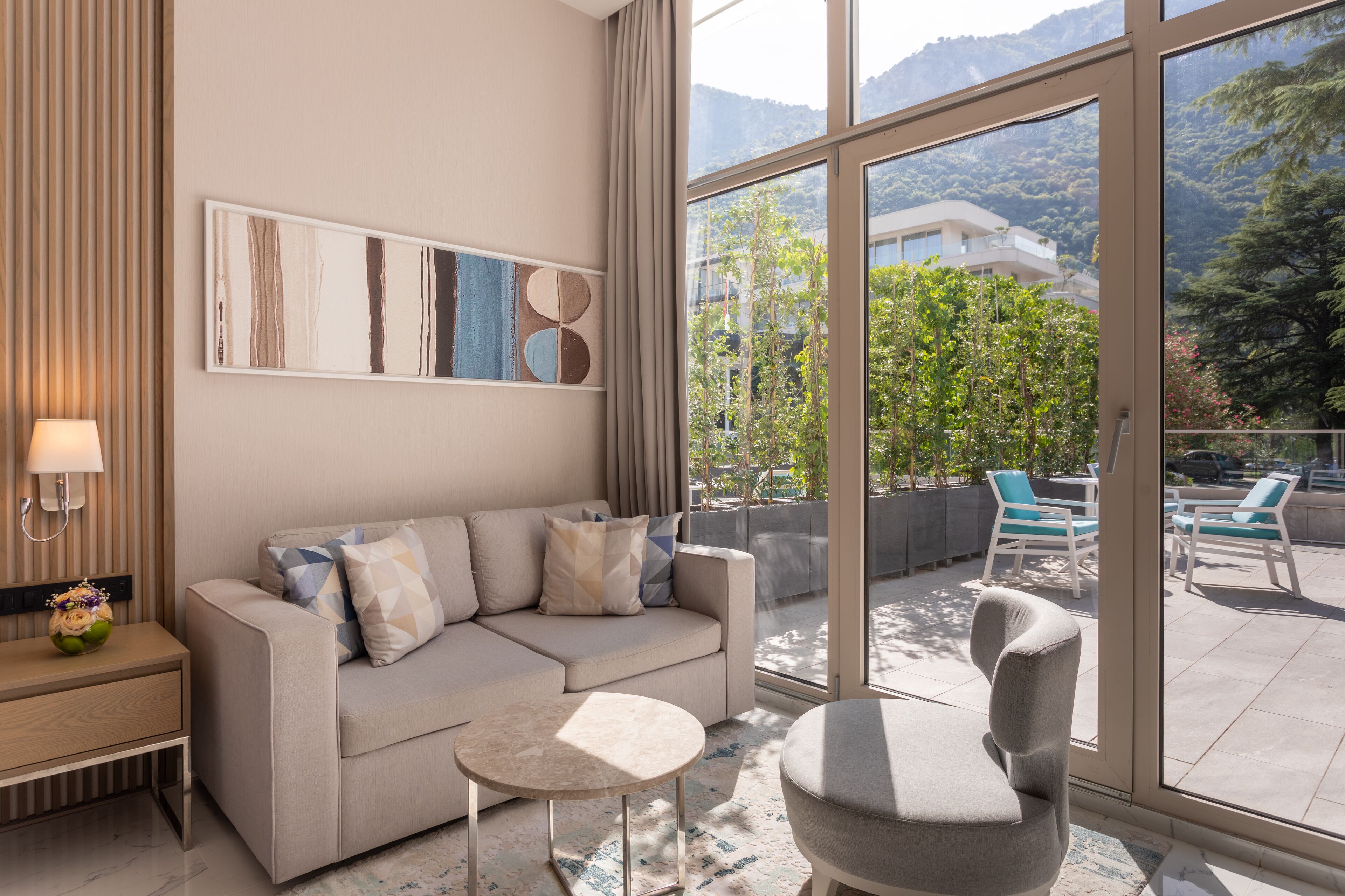 A modern living room with a sofa, a small round table, and a chair. There is a large window offering a view of a balcony with outdoor furniture.