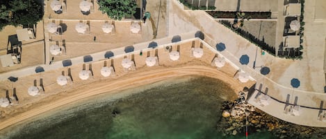 Vista aérea de una zona costera con playa arenosa, agua cristalina y un paseo marítimo con sombrillas y asientos.