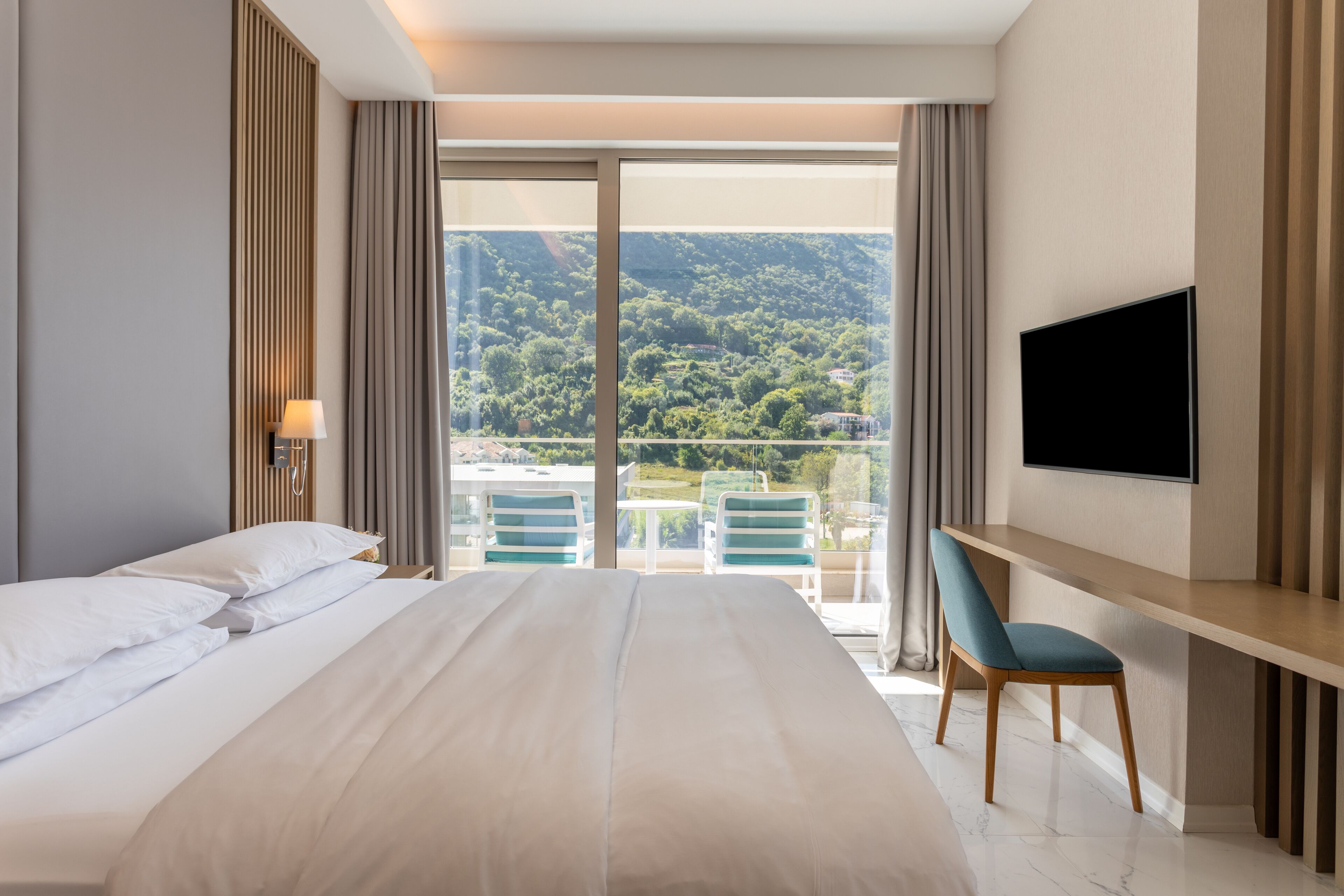 A hotel room with a large bed, a desk with a chair, a TV, and a balcony with a view of the mountains.