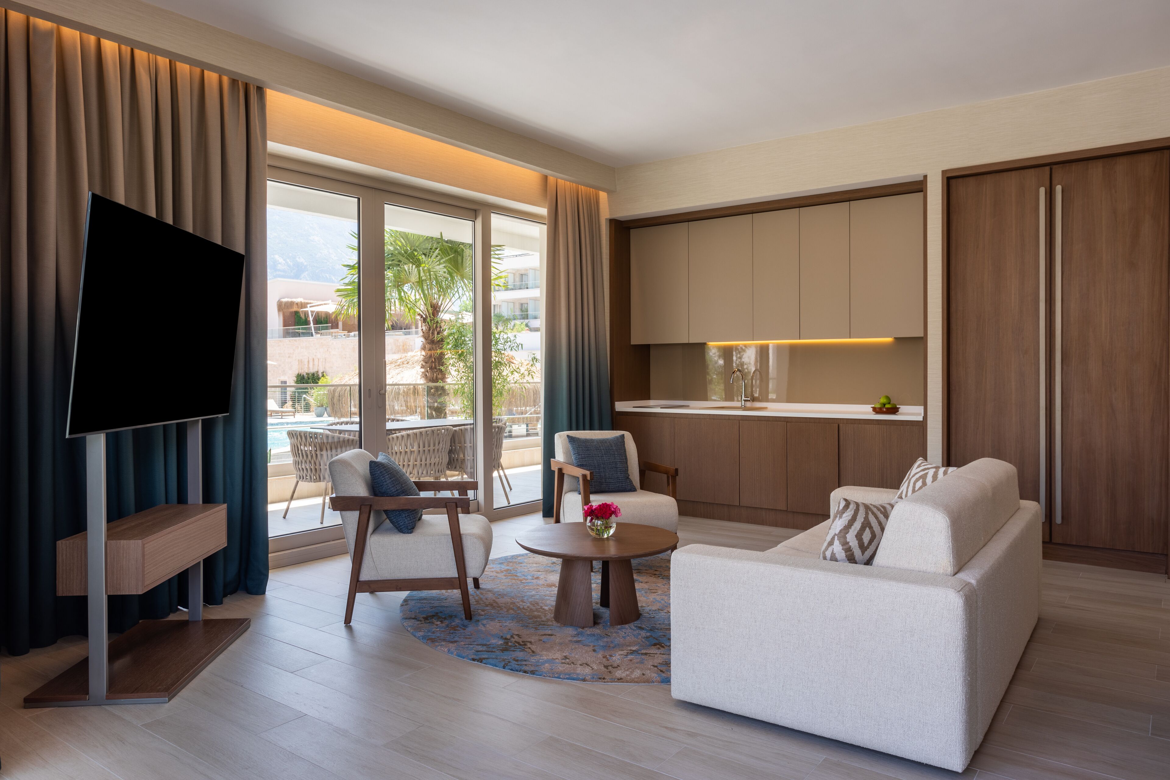 Une chambre d’hôtel moderne dotée d’une télévision à écran plat, d’un canapé, d’une table basse et offrant une vue sur une piscine et des palmiers.