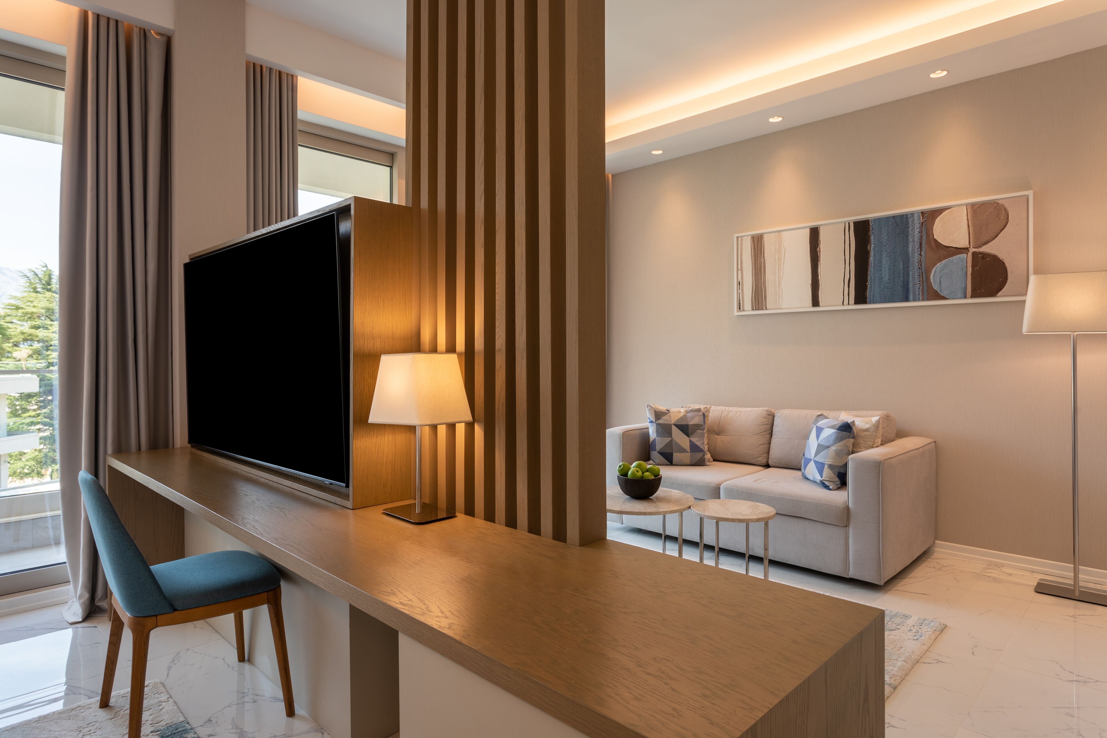 Un salon moderne avec un téléviseur à écran plat, un bureau en bois, une chaise bleue, un canapé avec des coussins à motifs et une lampe sur pied.