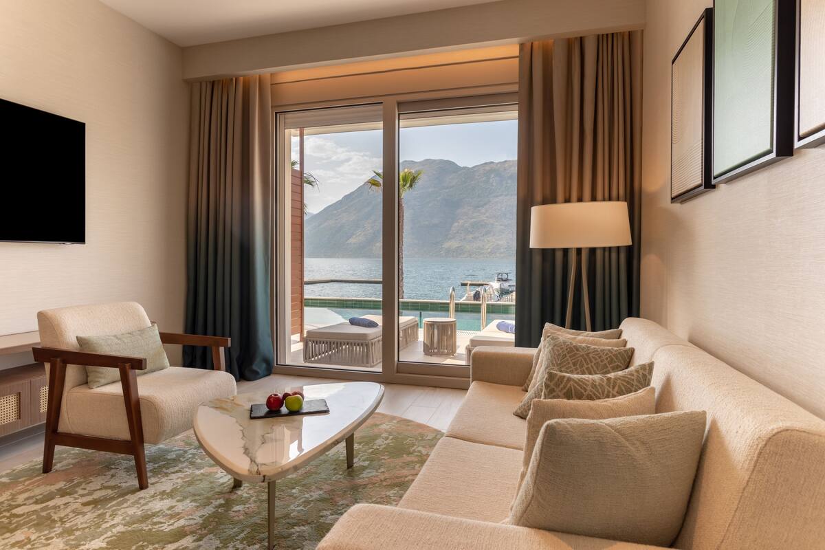 Une chambre d’hôtel avec un canapé, un fauteuil et une table basse. Un téléviseur est fixé au mur et une fenêtre offre une vue sur la mer et les montagnes.