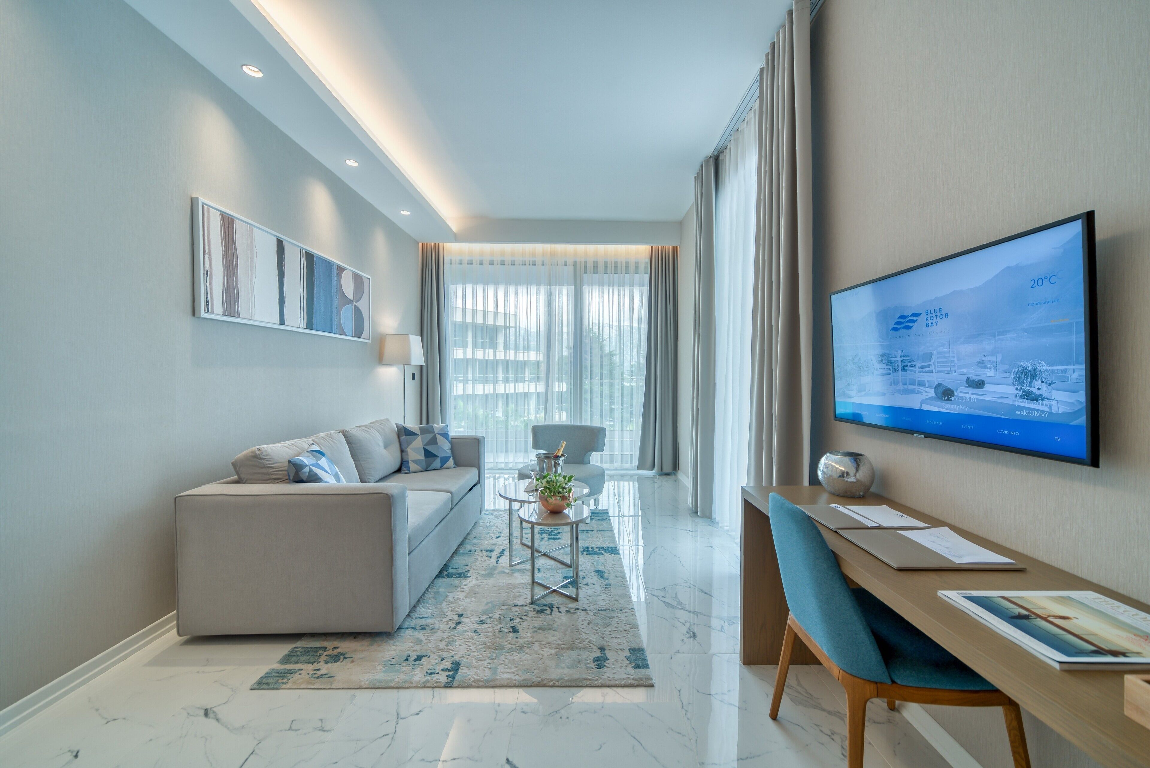 A modern hotel room with a sofa, a dining table, a TV, and a large window with curtains.
