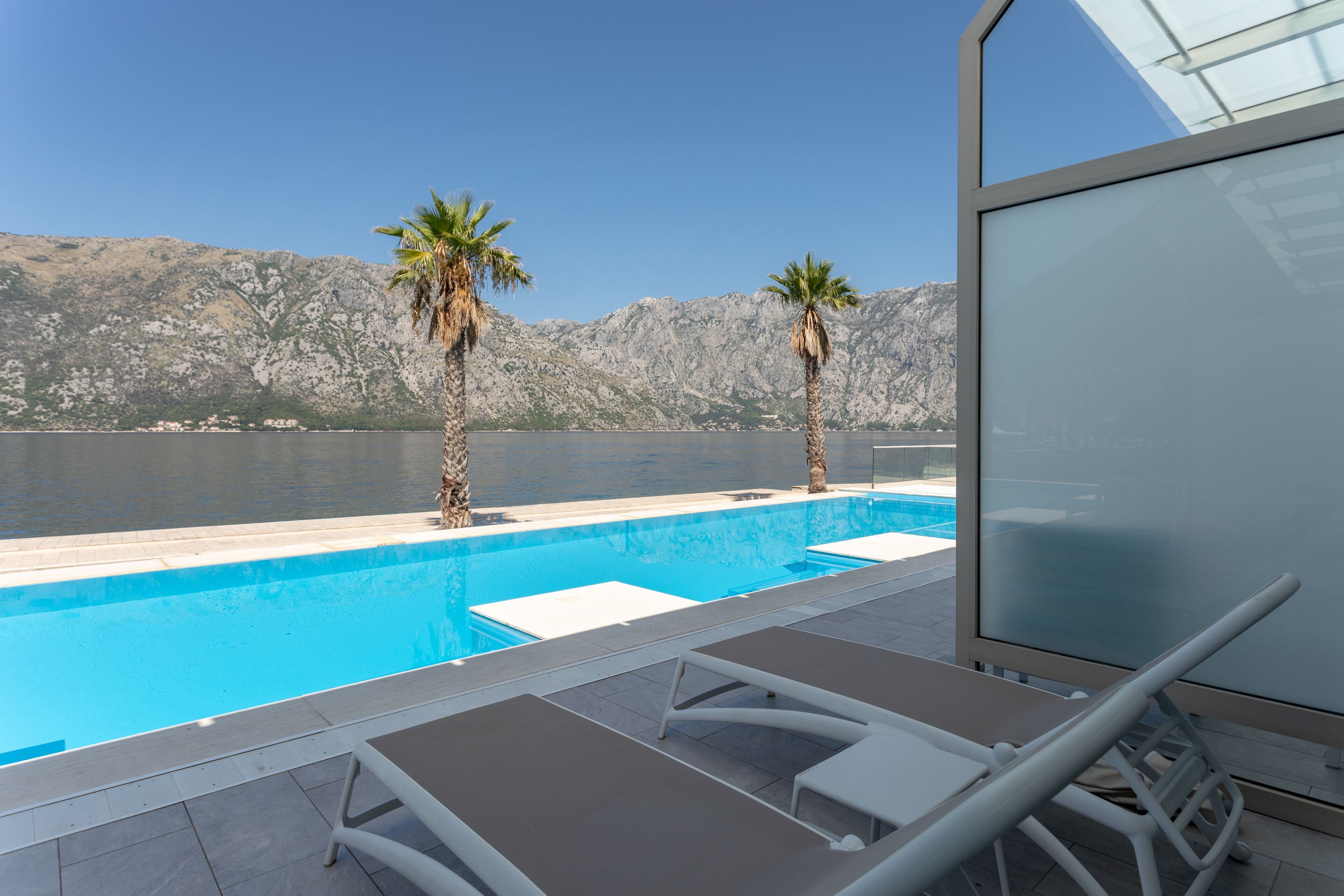 Una piscina con sillones de descanso y palmeras, con vistas a un cuerpo de agua y montañas.