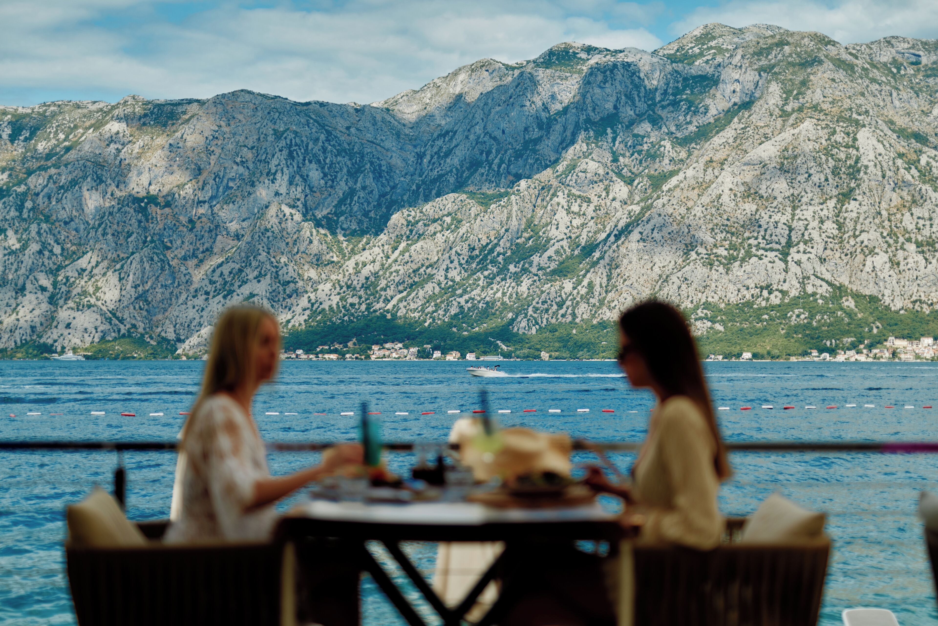 Deux personnes sont assises à une table, devant laquelle s’étend un paysage côtier montagneux.