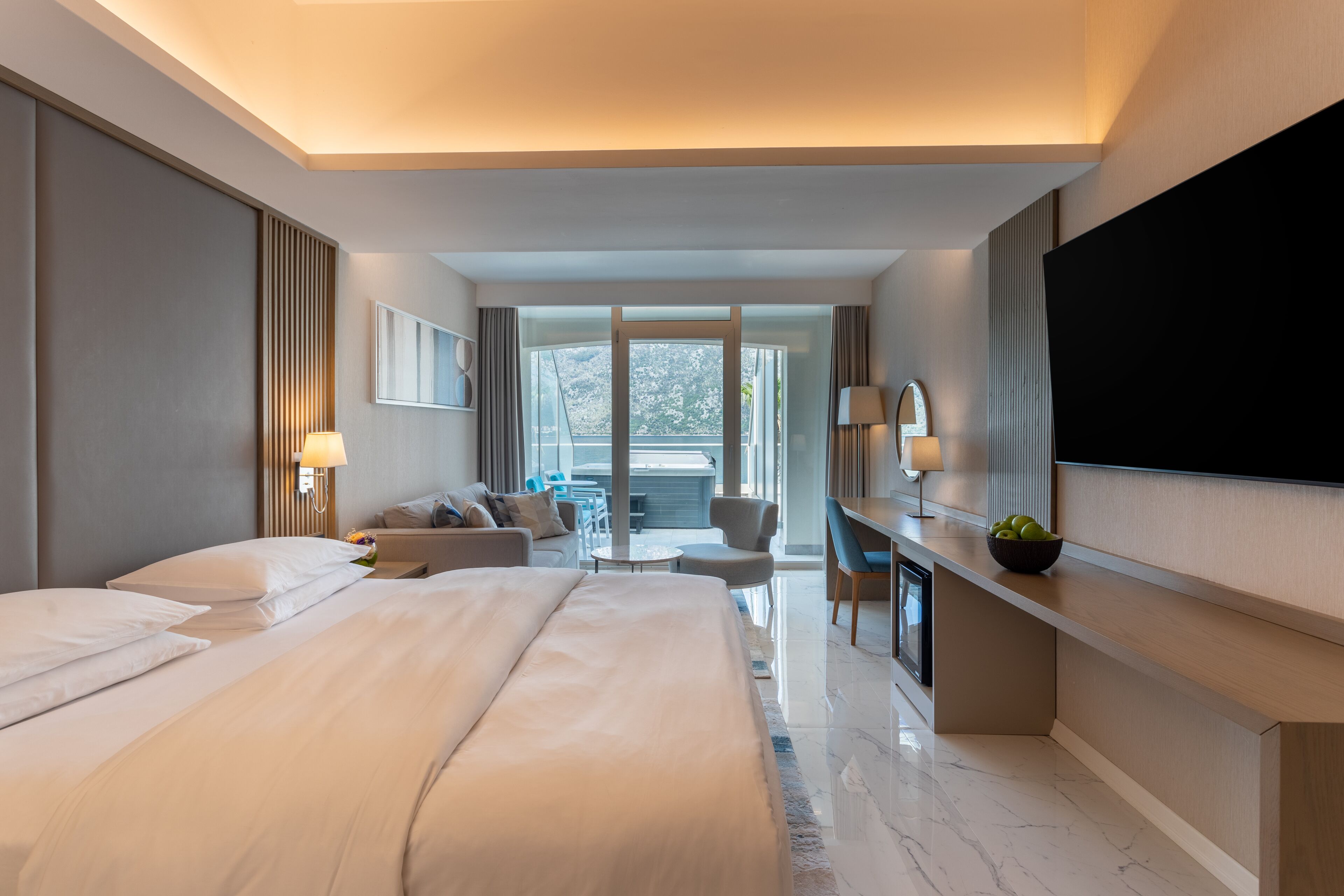 A modern hotel room with a large bed, a desk, a TV, and a balcony with a view of trees.
