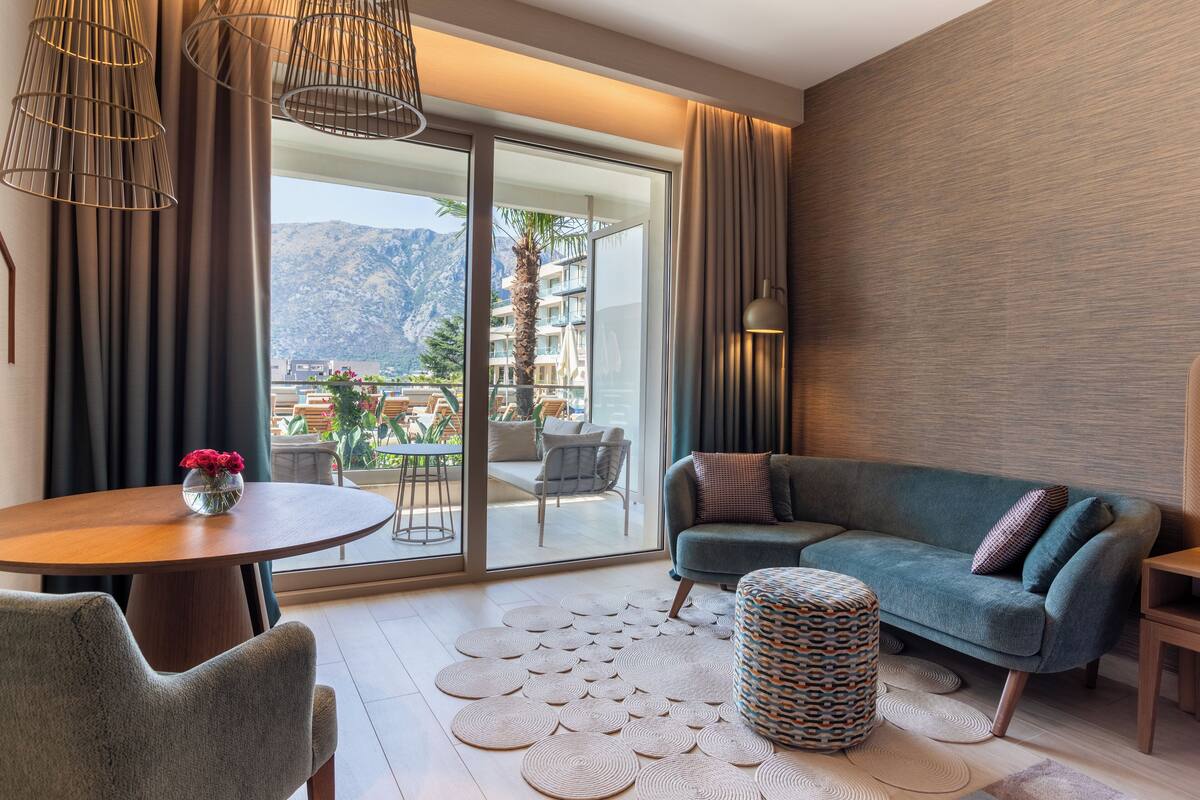 Un salon moderne avec un canapé, une table ronde et un pouf à motifs. Une grande fenêtre offre une vue sur un balcon aménagé avec du mobilier d’extérieur, et sur des montagnes en arrière-plan.