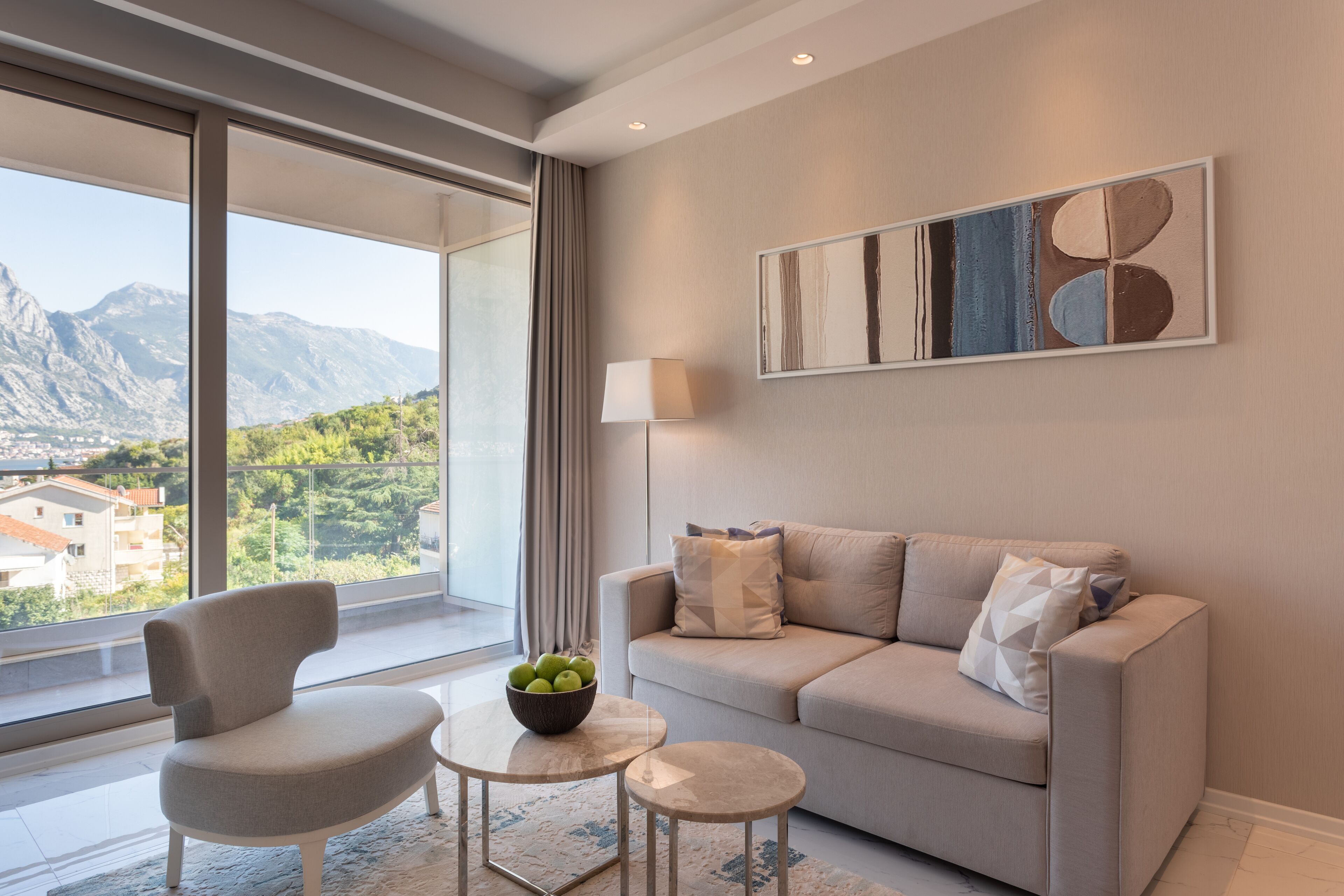Una sala de estar moderna con un sofá, un sillón y una mesita con un frutero. Un ventanal grande ofrece vistas a montañas y vegetación.