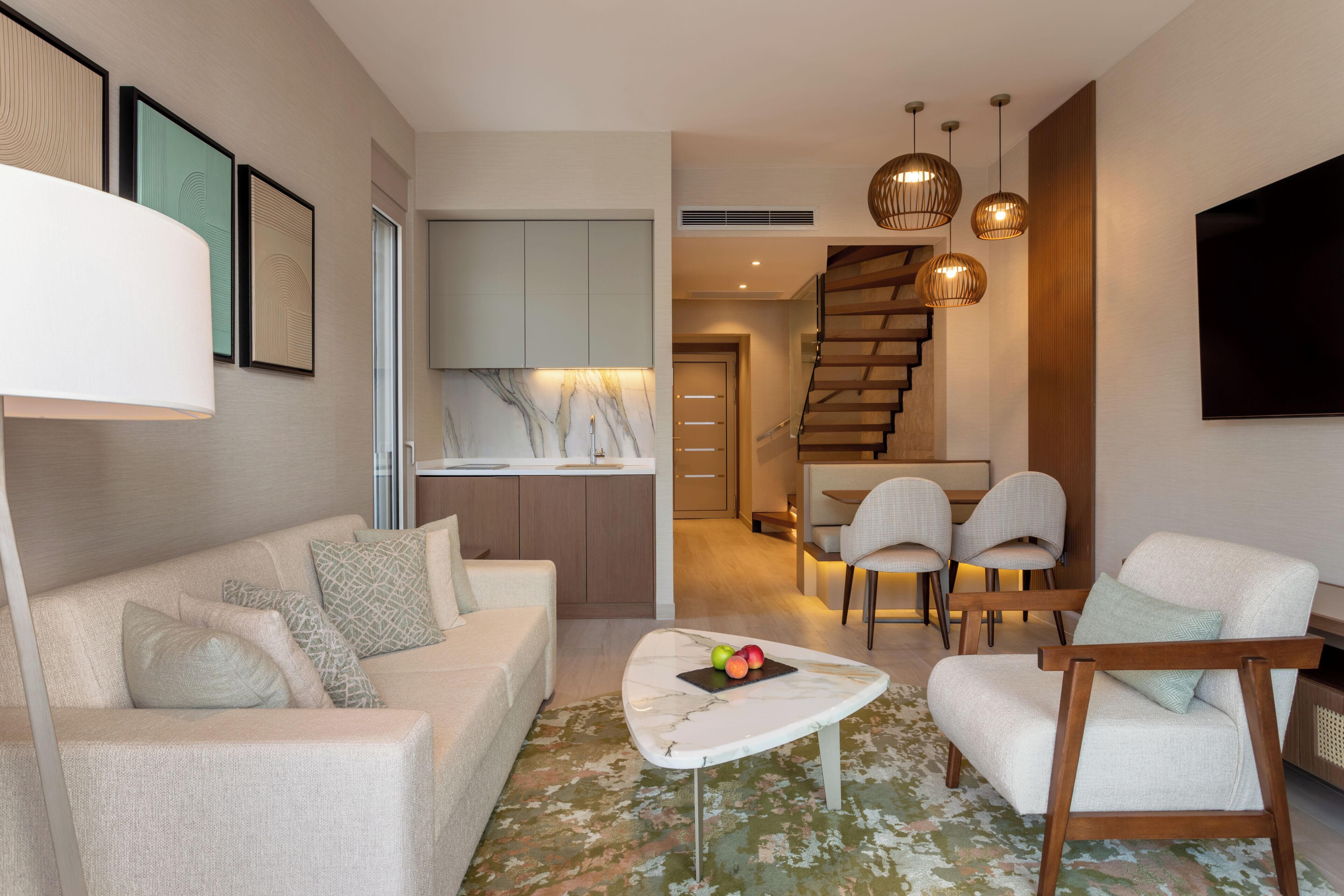Un salon moderne comprenant un canapé, des fauteuils, une table basse et un téléviseur. Un escalier mène à un espace repas avec des chaises.