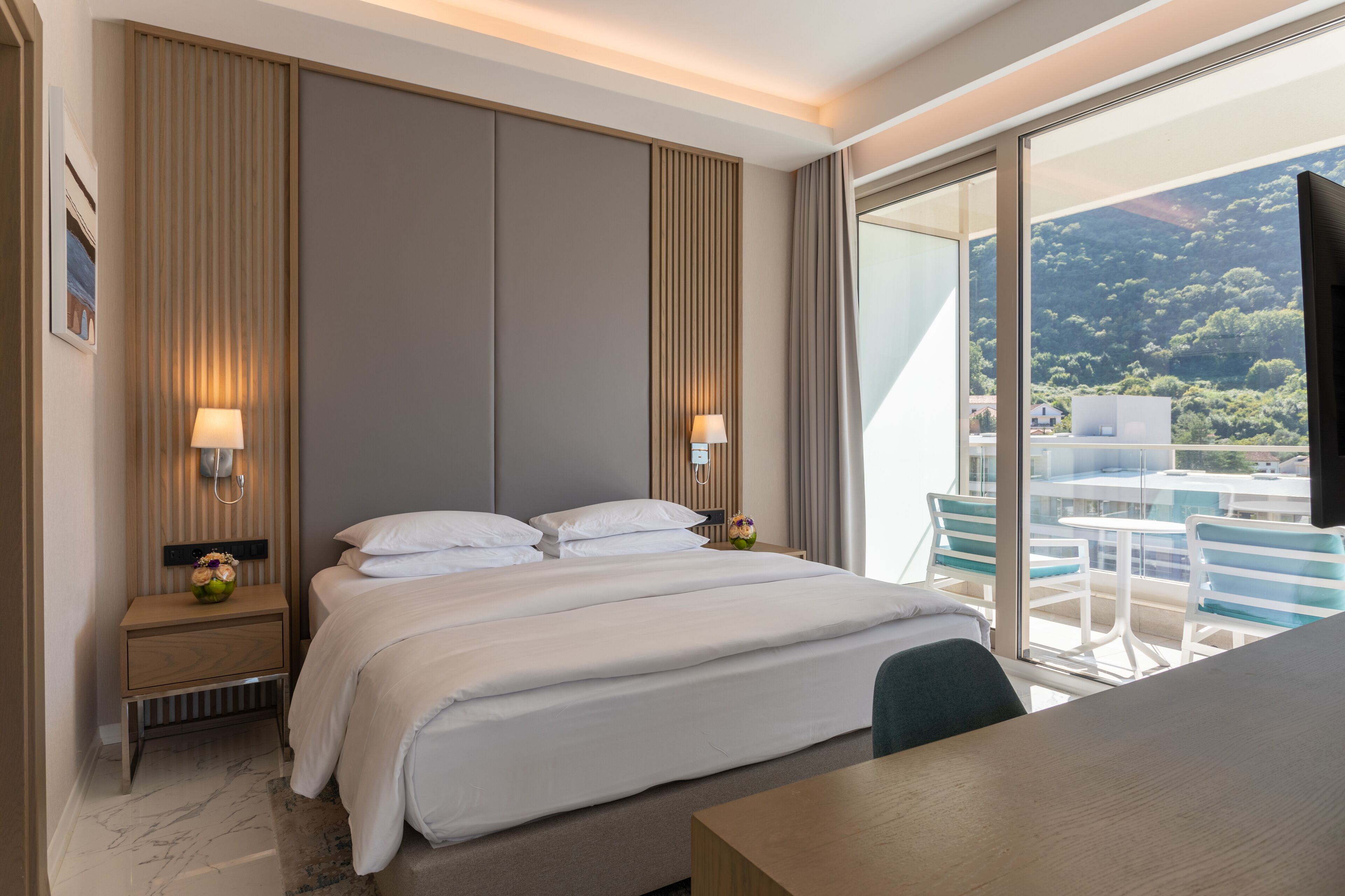 Quarto de hotel moderno com uma cama grande, luminárias de cabeceira, mesa de cabeceira de madeira e vista para uma varanda com cadeiras e uma mesa.