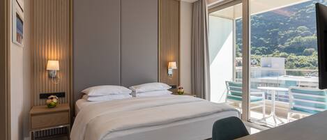 A modern hotel room with a large bed, bedside lamps, a wooden nightstand, and a view of a balcony with chairs and a table.