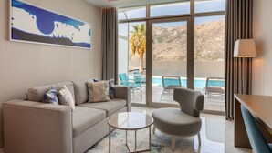 Habitación de hotel moderna con sofá, sillón y una pequeña mesa de centro. Amplias ventanas con vistas a una piscina y montañas.