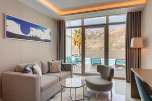 Habitación de hotel moderna con sofá, sillón y una pequeña mesa de centro. Amplias ventanas con vistas a una piscina y montañas.