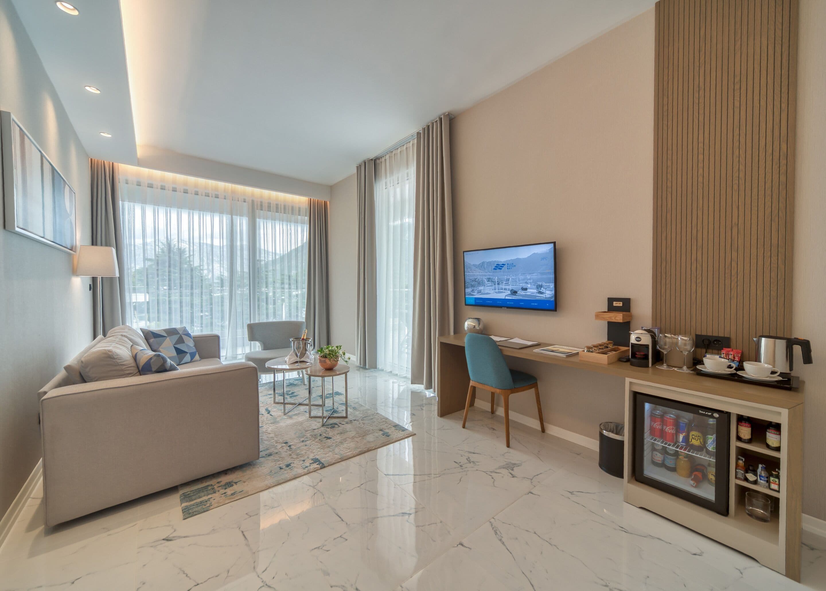 A modern hotel room with a sofa, a desk, a television, and a minibar.