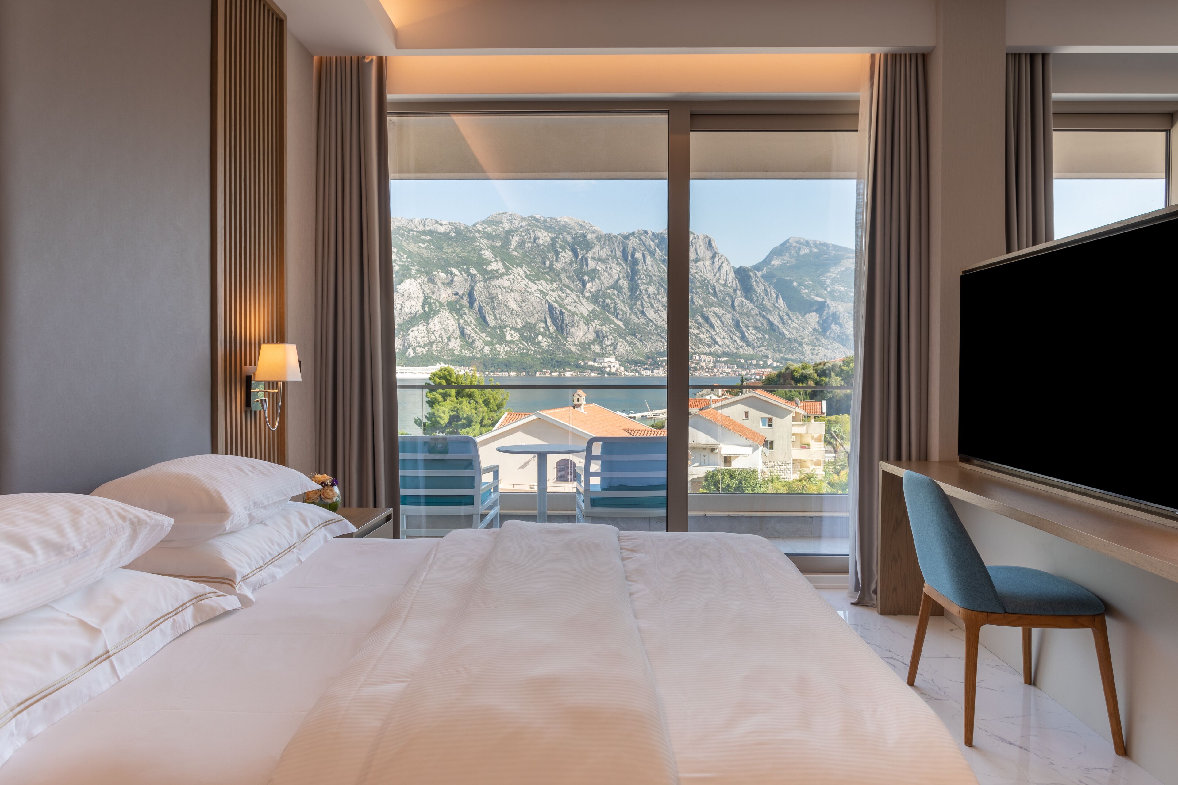 Habitación de hotel con una cama grande, un escritorio con una silla, televisión y vistas a montañas y un cuerpo de agua.