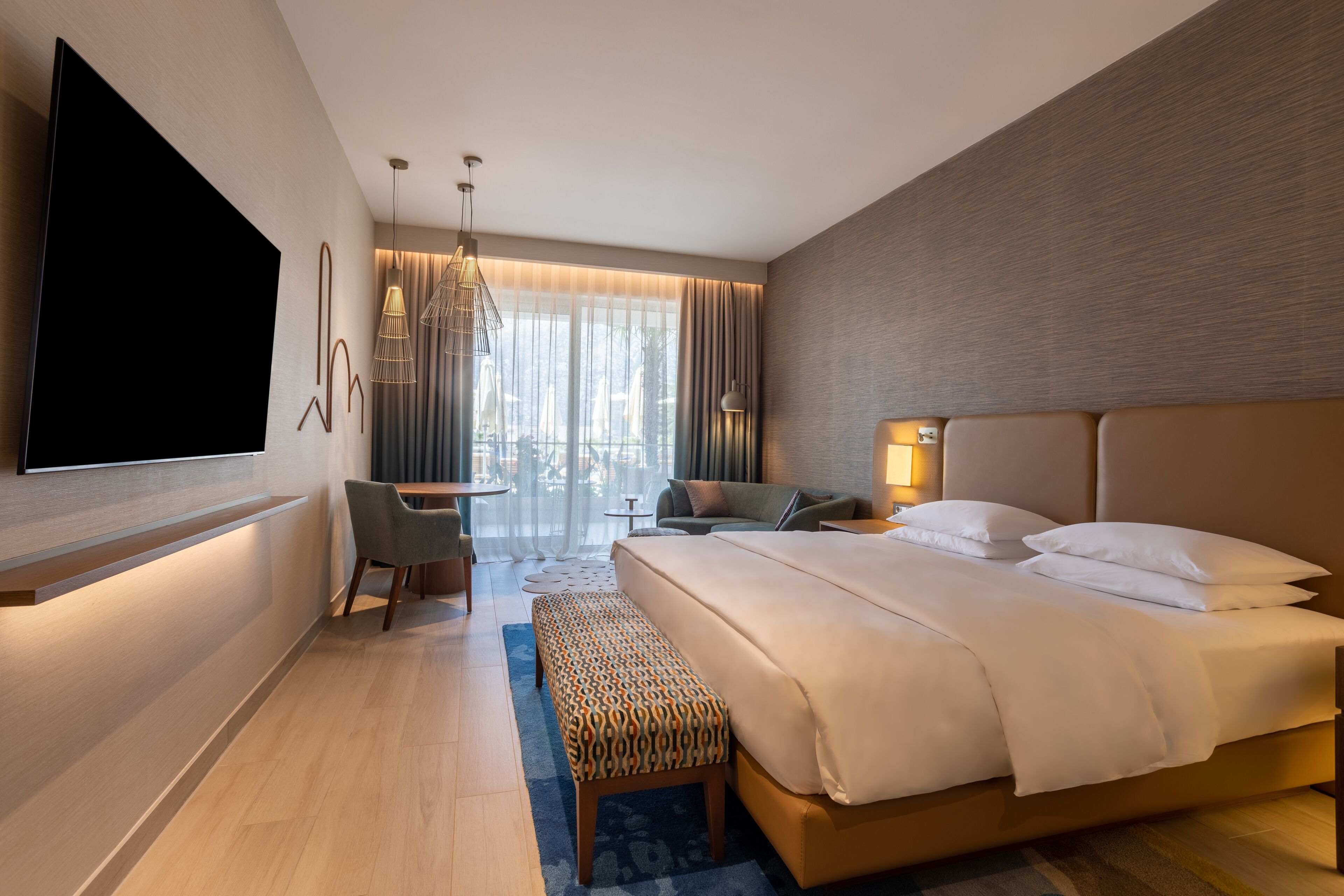 A modern hotel room with a large bed, a flat-screen TV mounted on the wall, a seating area with a chair and a small table, and a large window with curtains.