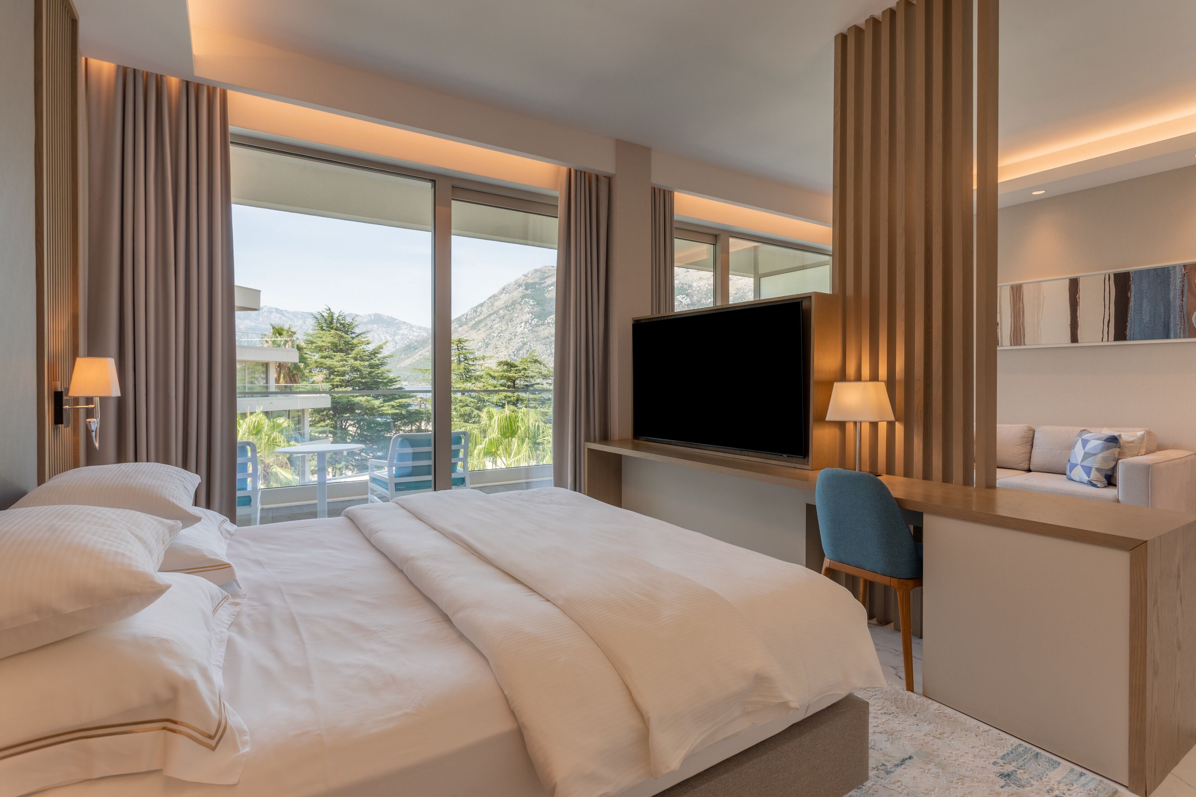 A hotel room with a large bed, a desk, a TV, and a balcony with a mountain view.