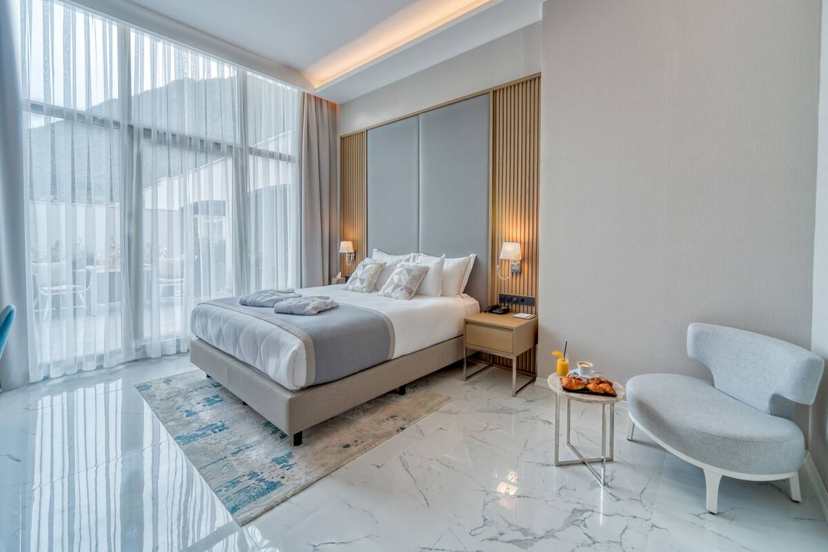 Une chambre d’hôtel moderne avec un grand lit, une table de chevet avec une lampe, une chaise et une vue sur l’extérieur.