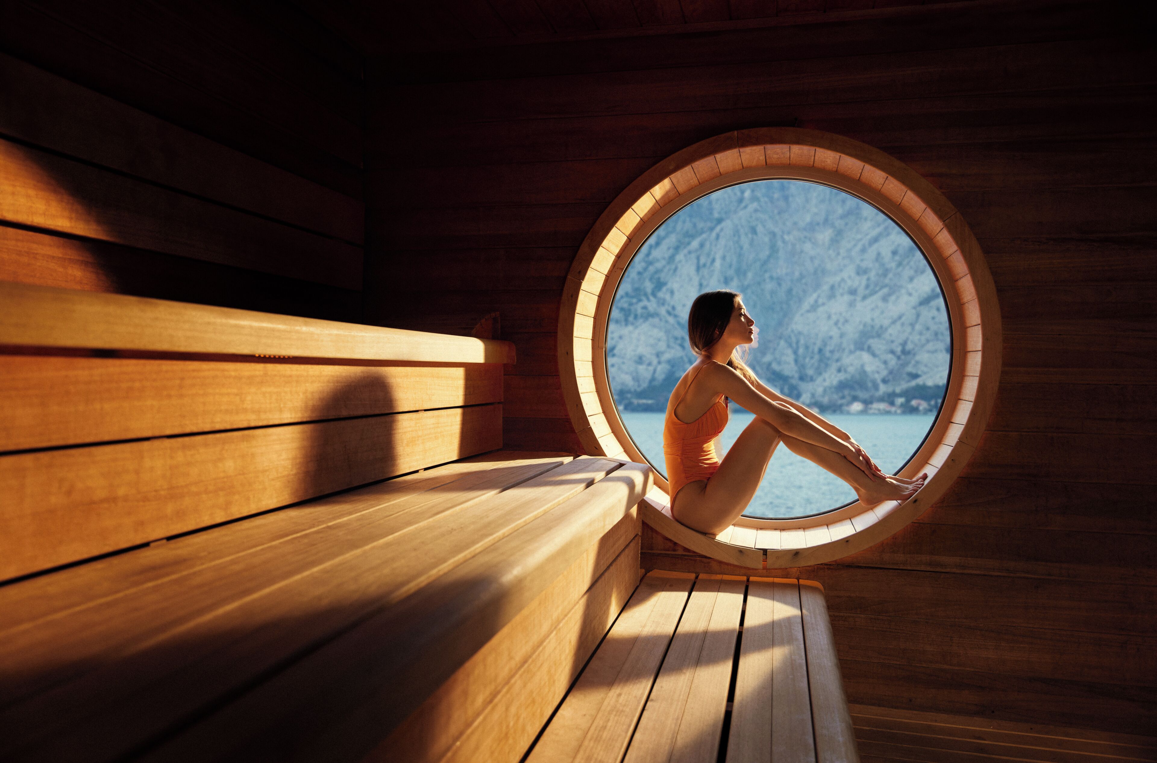 A person sitting by a circular window in a wooden room with a view of a mountainous landscape.