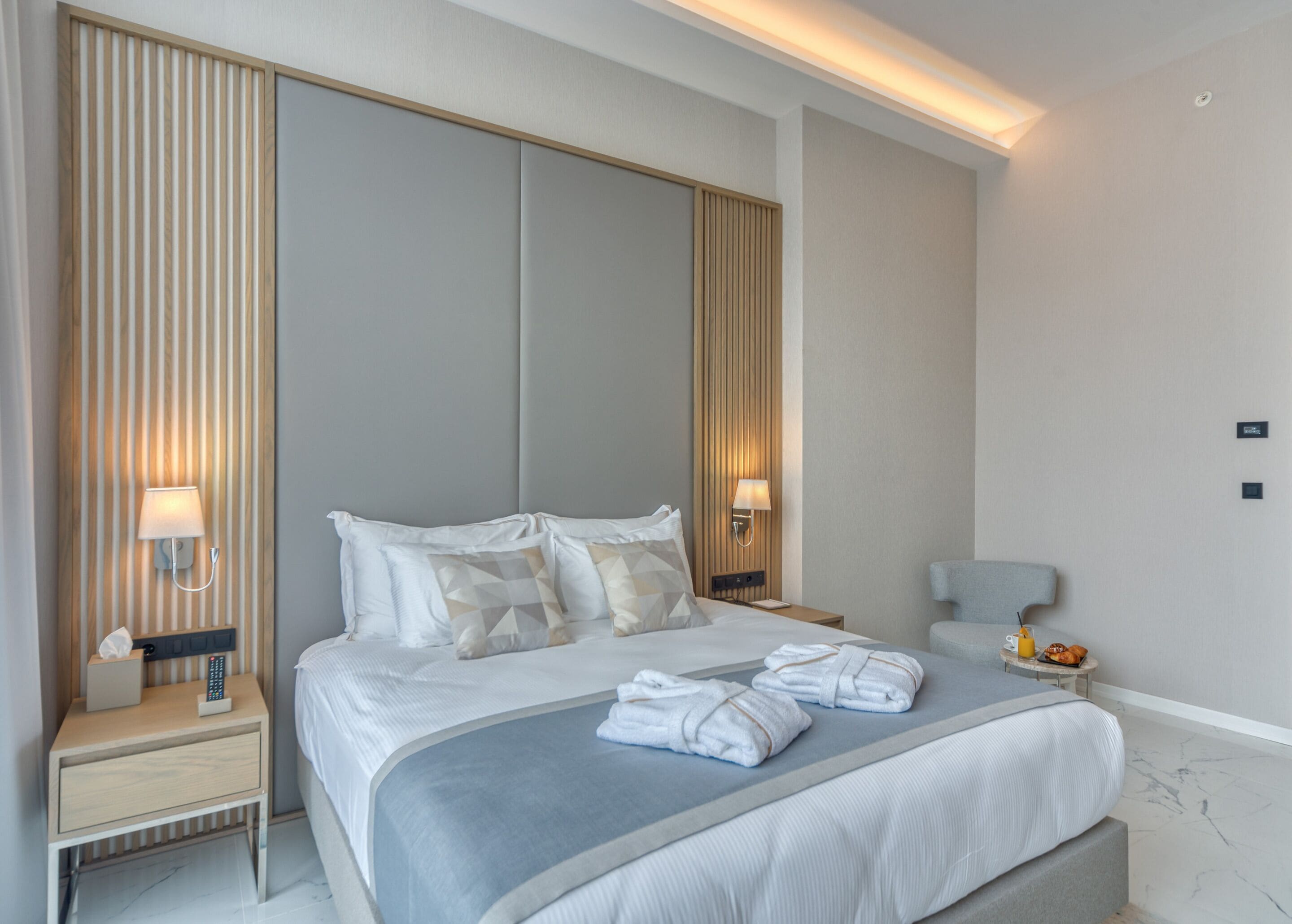A modern hotel room with a large bed, bedside tables, a nightstand, a chair, and a small table with a fruit bowl.