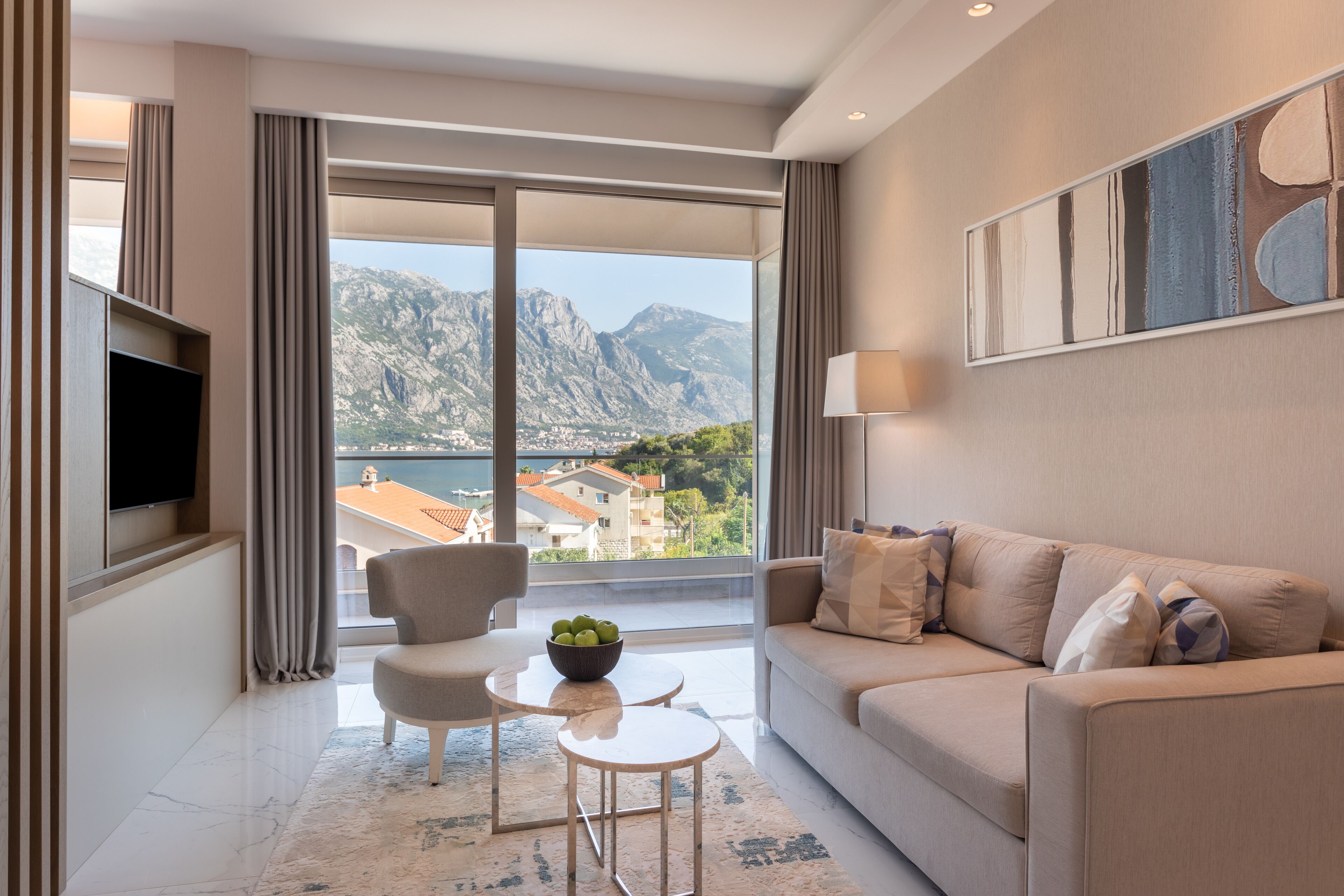 A modern living room with a sofa, armchairs, a small table with fruit, and a TV. A large window offers a view of mountains and a lake.